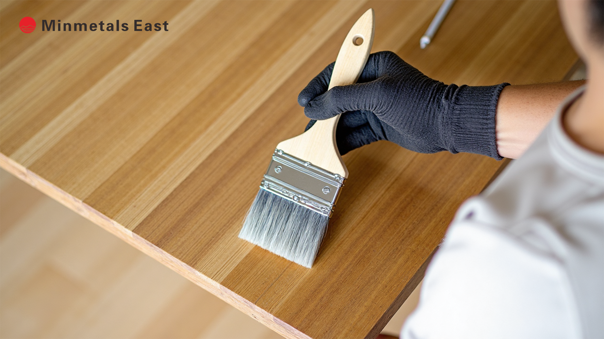 A person wearing a black glove holds a paintbrush with a wooden handle and light-colored bristles, applying a finish to a light-toned, striped wooden surface, possibly a tabletop. The 'Minmetals East' logo is visible in the top left corner.